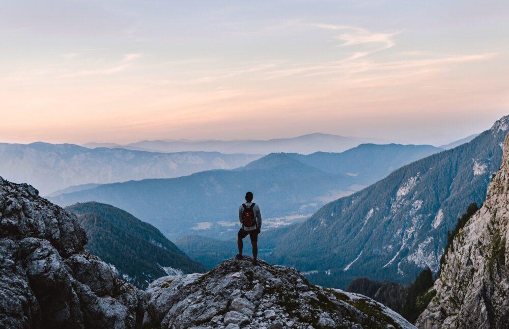 A person with a backpack stands on a rocky peak, overlooking a scenic valley and distant mountains at sunrise or sunset, with mist and layers of hills fading into the horizon under a colorful sky.