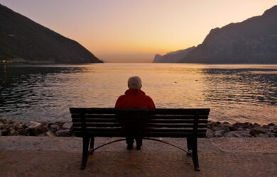 An elderly person sitting alone on a bench by a lake, facing calm water and distant mountains at sunset, with soft orange and purple hues in the sky.