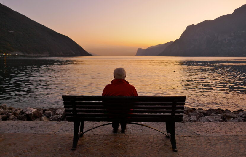 An elderly person sitting alone on a bench by a lake, facing calm water and distant mountains at sunset, with soft orange and purple hues in the sky.