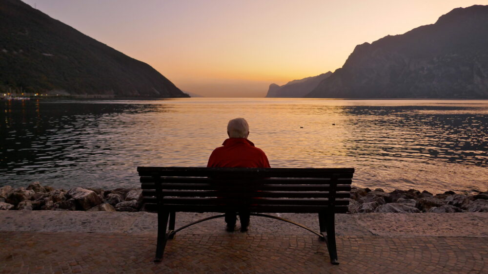 An elderly person sitting alone on a bench by a lake, facing calm water and distant mountains at sunset, with soft orange and purple hues in the sky.