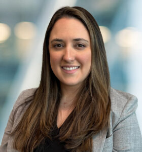 A woman with long brown hair, wearing a gray blazer and black top, smiles at the camera with a blurred background of lights and blue tones.