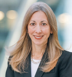 A woman with long, straight, light brown hair wearing a black blazer, white top, and a beaded necklace, smiles at the camera with a blurred modern office background.