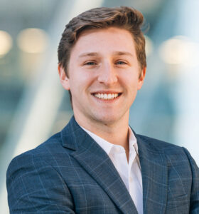 A young man in a checkered suit jacket and white shirt smiles confidently while standing with arms crossed against a blurred, modern glass building background.