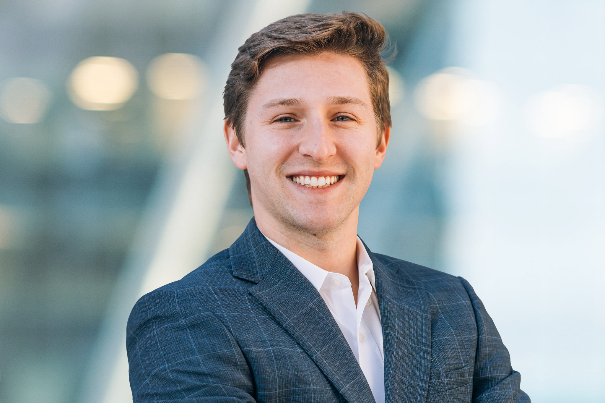 A young man in a checkered suit jacket and white shirt smiles confidently while standing with arms crossed against a blurred, modern glass building background.
