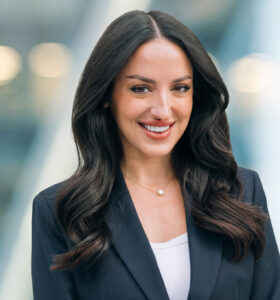 A woman with long dark hair wearing a dark blazer and white top smiles confidently at the camera, standing in front of a blurred modern office background.