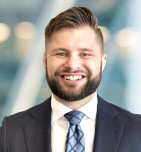 A smiling man with short brown hair and a full beard wears a navy suit, white shirt, and patterned tie, standing in front of a blurred office background with soft lighting.