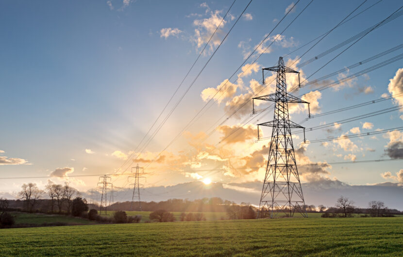 High-voltage power lines and pylons stretch across a green field at sunset, with sunlight shining through scattered clouds in the sky. Trees are visible in the background.