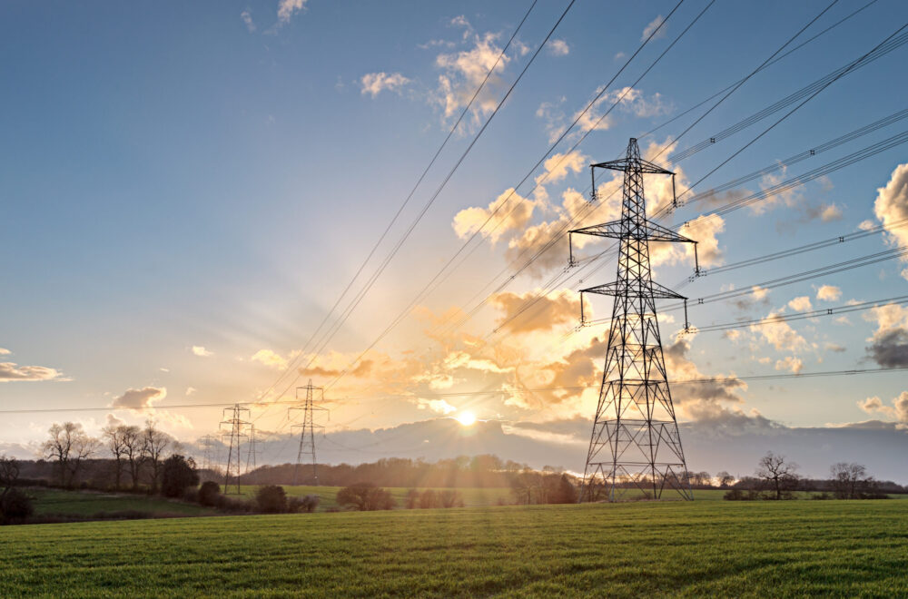 High-voltage power lines and pylons stretch across a green field at sunset, with sunlight shining through scattered clouds in the sky. Trees are visible in the background.