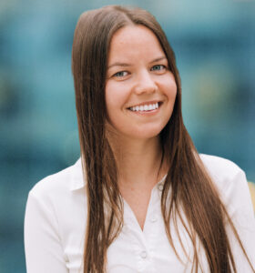 A young woman with long brown hair smiles while wearing a white button-up shirt, standing in front of a blurred blue and yellow background.