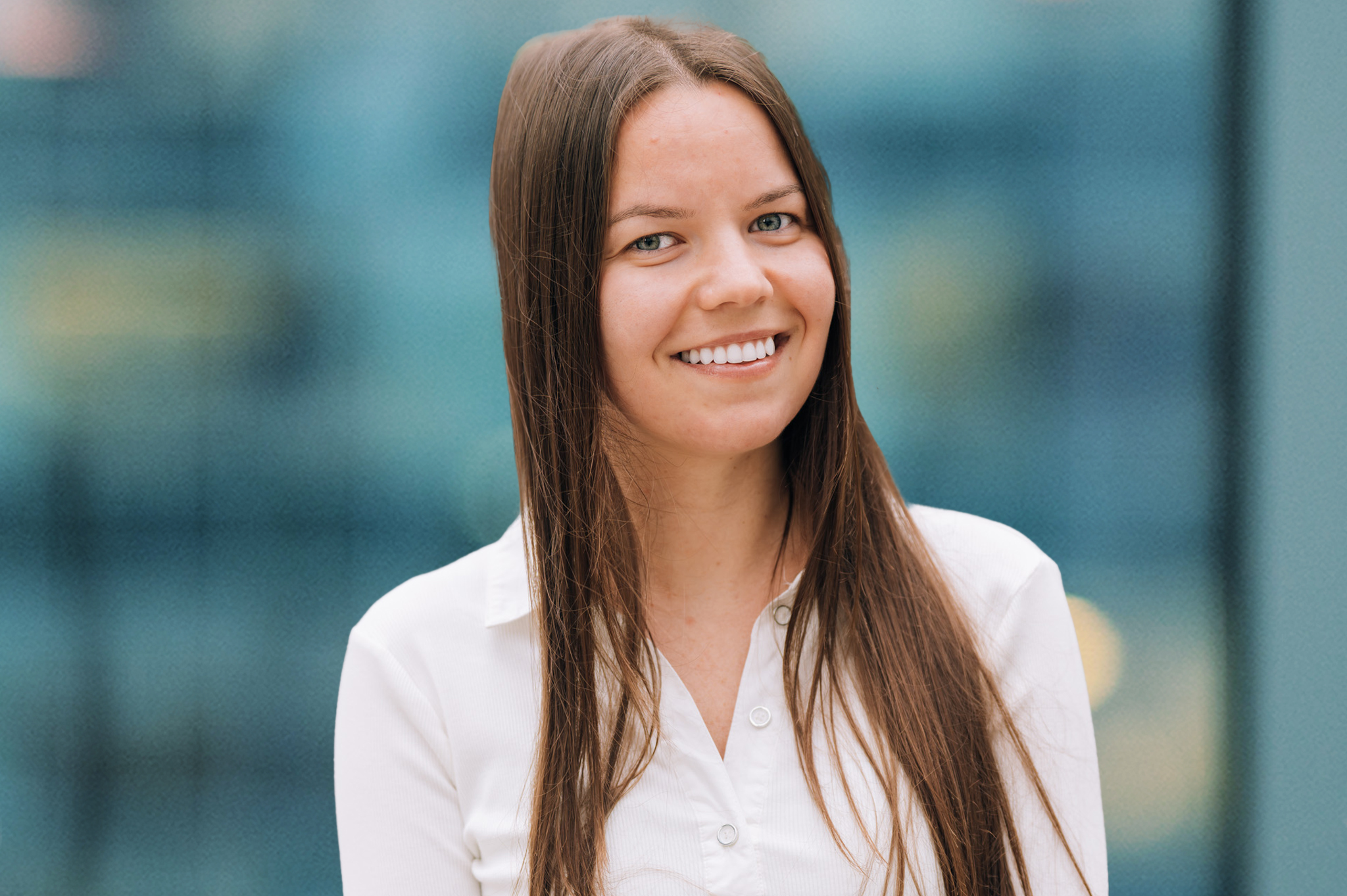 A young woman with long brown hair smiles while wearing a white button-up shirt, standing in front of a blurred blue and yellow background.