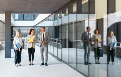 Three business professionals, two women and one man, stand and talk outside an office building with large glass windows, holding folders and documents. Their reflections are visible in the glass.