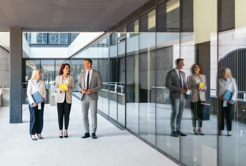 Three business professionals, two women and one man, stand and talk outside an office building with large glass windows, holding folders and documents. Their reflections are visible in the glass.