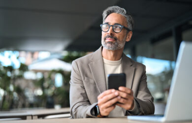 A middle-aged man with gray hair and a beard, wearing glasses and a beige blazer, sits at a table with a laptop, holding a smartphone and looking thoughtfully into the distance.