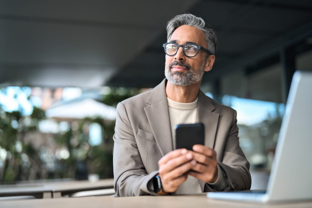 A middle-aged man with gray hair and a beard, wearing glasses and a beige blazer, sits at a table with a laptop, holding a smartphone and looking thoughtfully into the distance.