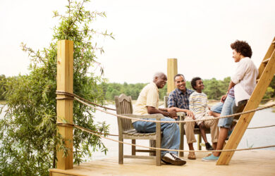A multigenerational family sits and talks together on a wooden dock by a lake, surrounded by greenery, enjoying a relaxed and happy moment outdoors.