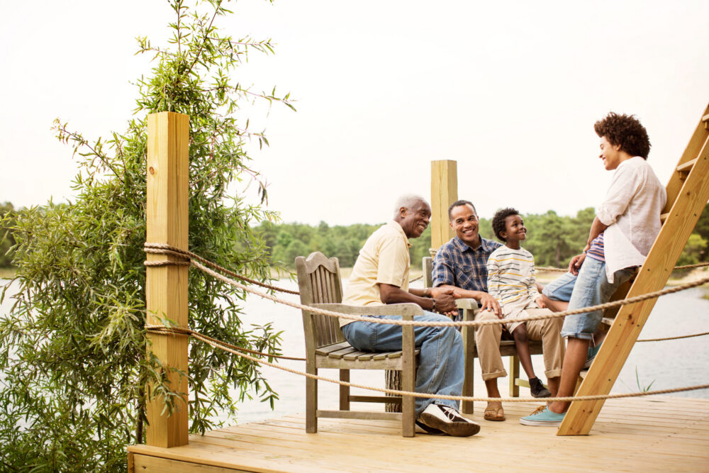 A multigenerational family sits and talks together on a wooden dock by a lake, surrounded by greenery, enjoying a relaxed and happy moment outdoors.