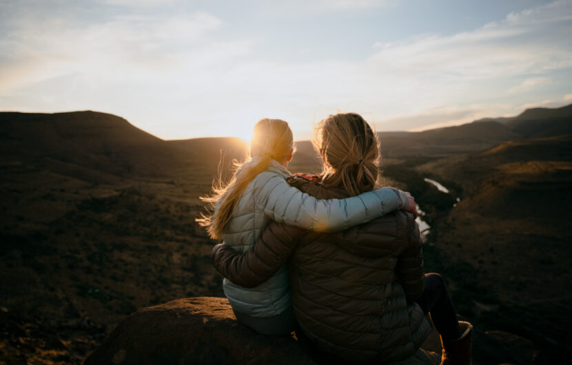 Two people in jackets sit on a rock with arms around each other, watching the sunset over a vast, open landscape with hills and a winding river.