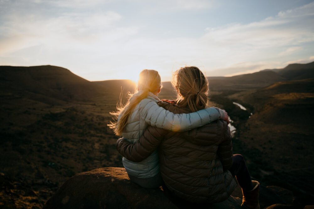Two people in jackets sit on a rock with arms around each other, watching the sunset over a vast, open landscape with hills and a winding river.