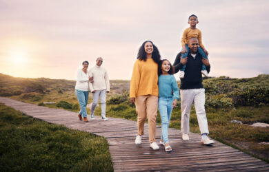 A multi-generational family walks along a wooden path outdoors at sunset, with parents, children, and grandparents smiling together amid grassy scenery.
