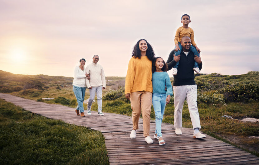 A multi-generational family walks along a wooden path outdoors at sunset, with parents, children, and grandparents smiling together amid grassy scenery.