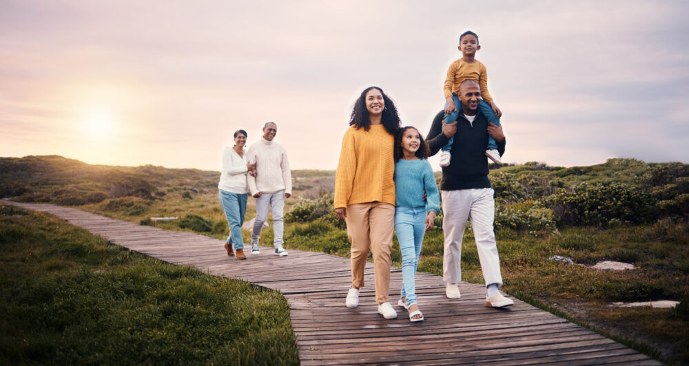 A multi-generational family walks along a wooden path outdoors at sunset, with parents, children, and grandparents smiling together amid grassy scenery.