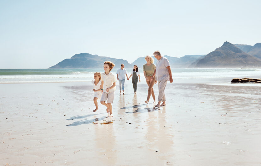 Three generations of a family walk on a sandy beach by the ocean; two children run ahead while adults follow behind, holding hands, with mountains and blue sky in the background.