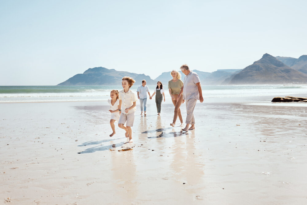 Three generations of a family walk on a sandy beach by the ocean; two children run ahead while adults follow behind, holding hands, with mountains and blue sky in the background.