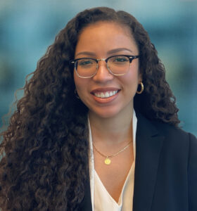 A woman with long curly hair, wearing glasses, a black blazer, white blouse, and gold jewelry, smiles at the camera. The background is softly blurred, suggesting an office setting.