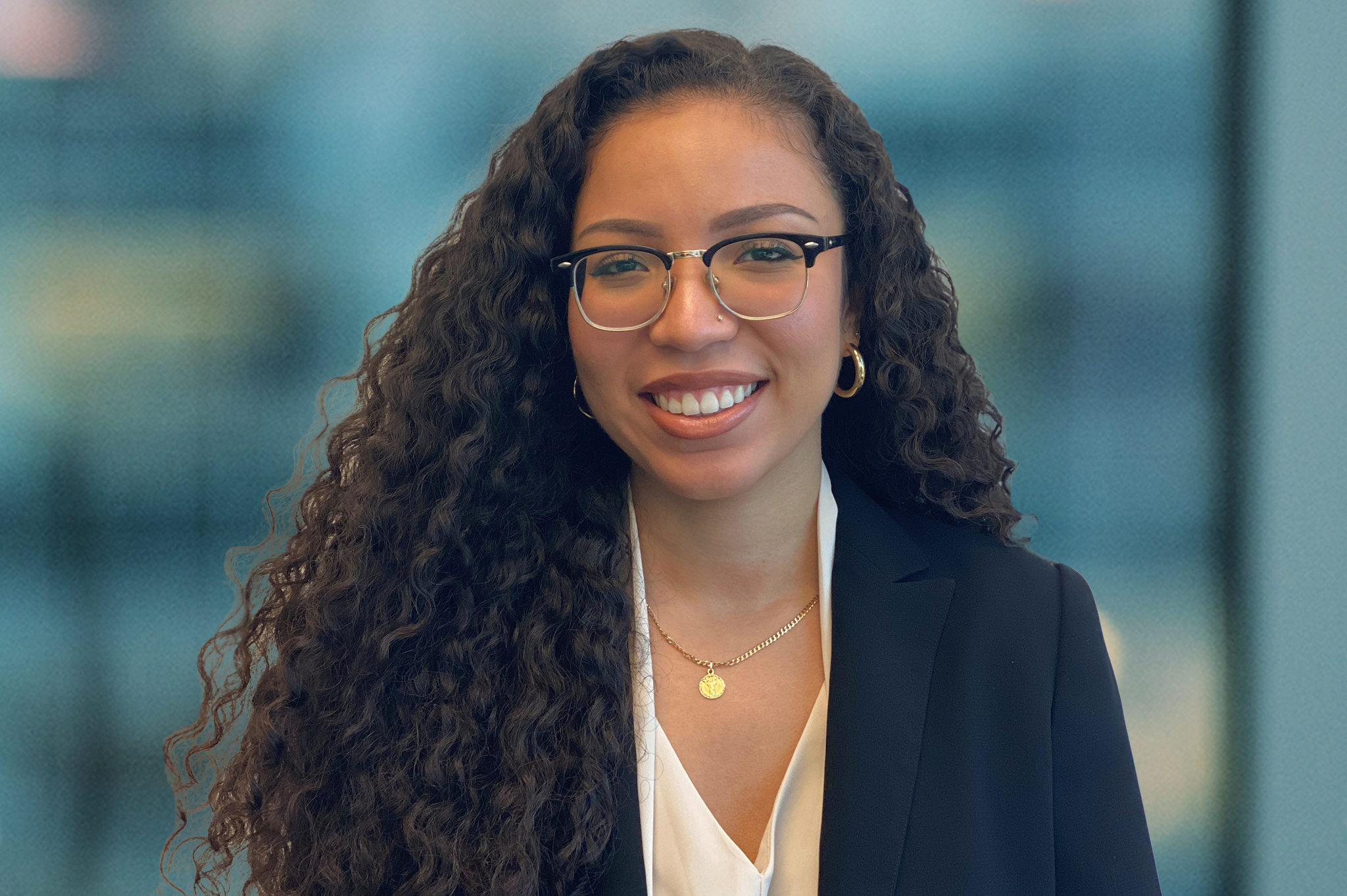A woman with long curly hair, wearing glasses, a black blazer, white blouse, and gold jewelry, smiles at the camera. The background is softly blurred, suggesting an office setting.