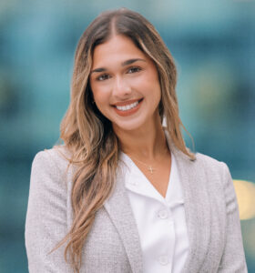 A woman with long, wavy hair wearing a light gray blazer and white blouse smiles at the camera with a blurred blue background behind her.