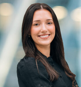 A woman with long dark hair, wearing a black blouse, stands smiling with arms crossed in front of a blurred background with soft lights.