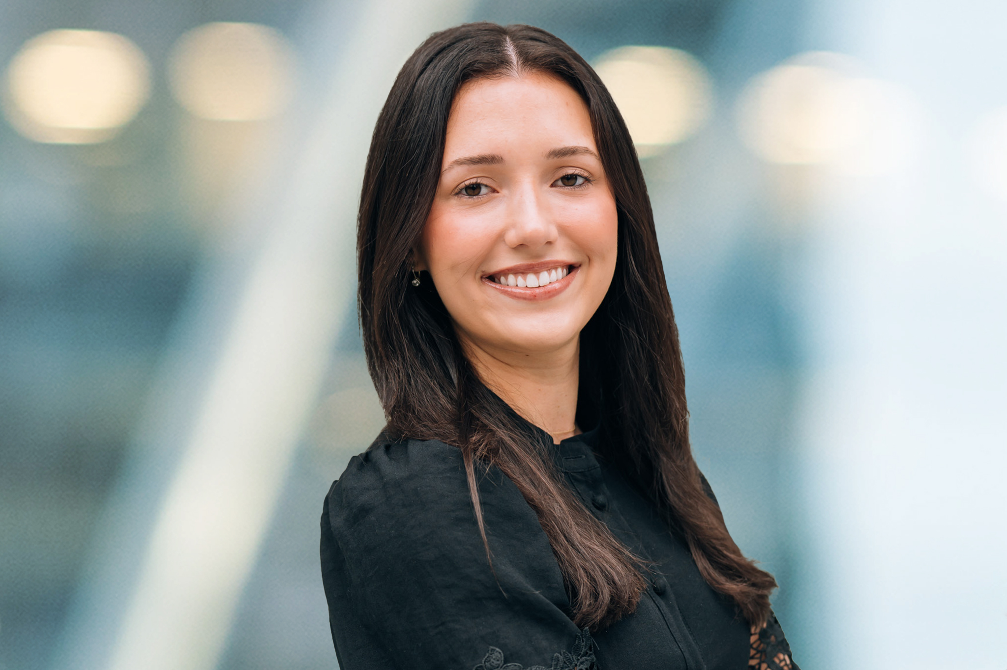 A woman with long dark hair, wearing a black blouse, stands smiling with arms crossed in front of a blurred background with soft lights.