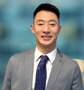 A young man wearing a gray suit, white dress shirt, and patterned blue tie stands smiling in front of a blurred blue background.