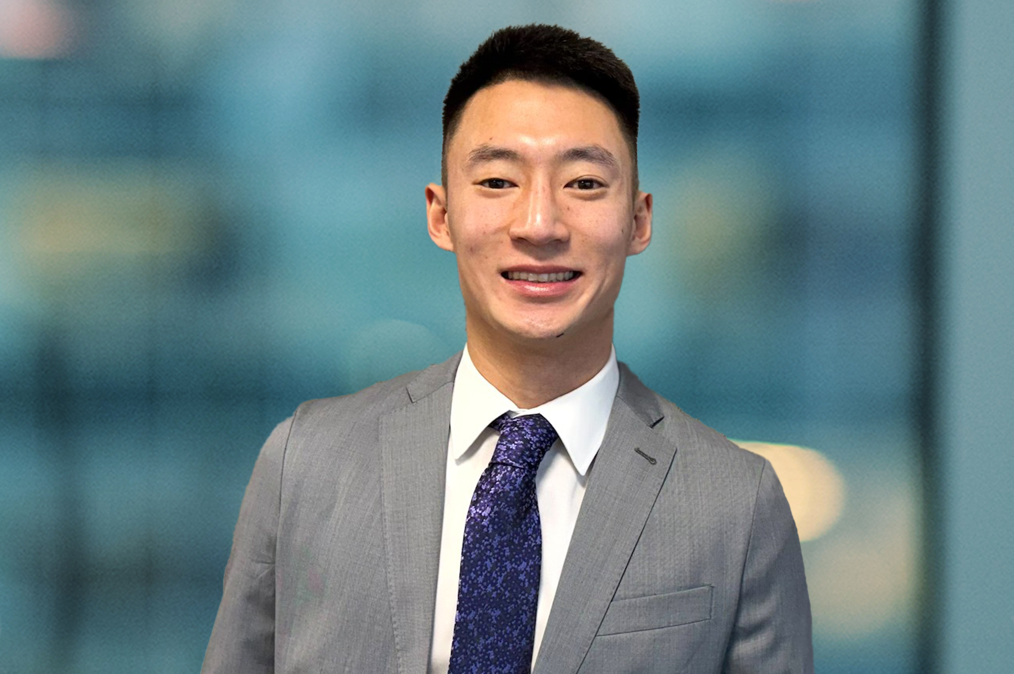 A young man wearing a gray suit, white dress shirt, and patterned blue tie stands smiling in front of a blurred blue background.