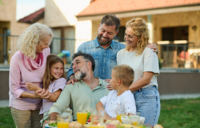 Three generations of a family smile and gather around a picnic table outdoors, enjoying each other’s company and a meal with drinks and food on a sunny day.