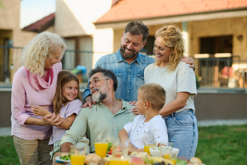 Three generations of a family smile and gather around a picnic table outdoors, enjoying each other’s company and a meal with drinks and food on a sunny day.