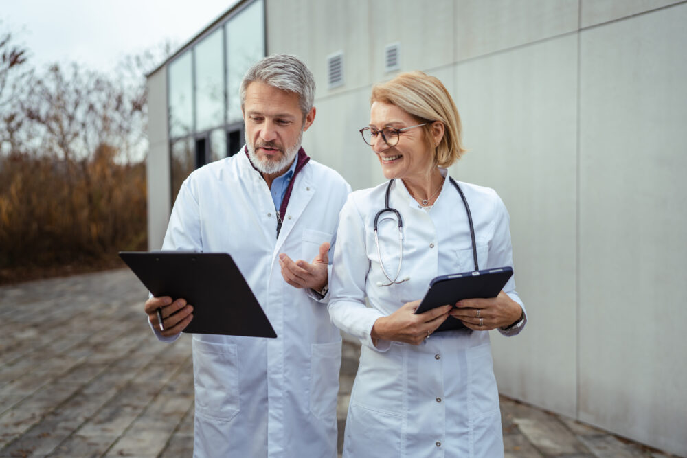 Two doctors in white coats, one with a stethoscope, stand outside a building, smiling and discussing documents on clipboards.