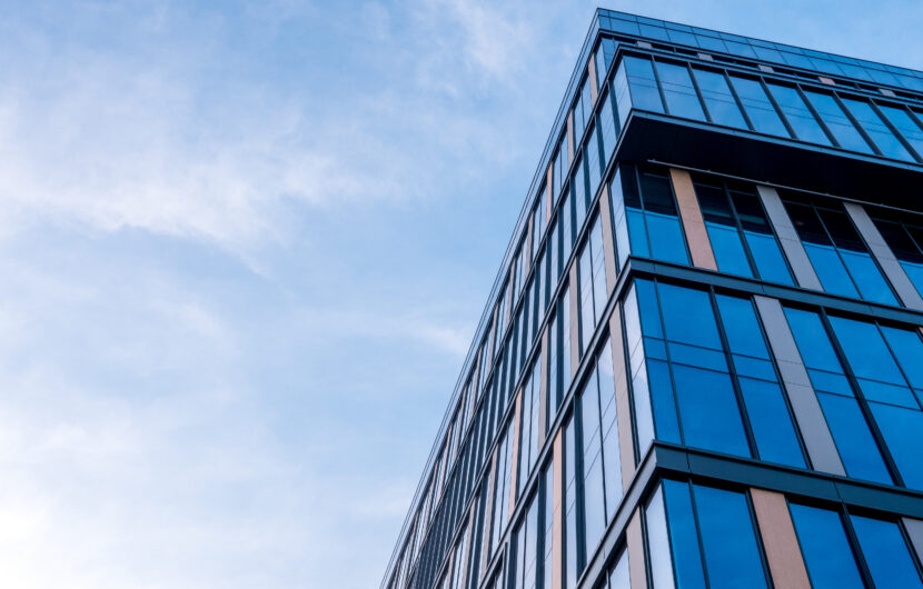Modern glass office building viewed from a low angle against a blue sky with light clouds, showing reflective windows and sleek architectural lines.