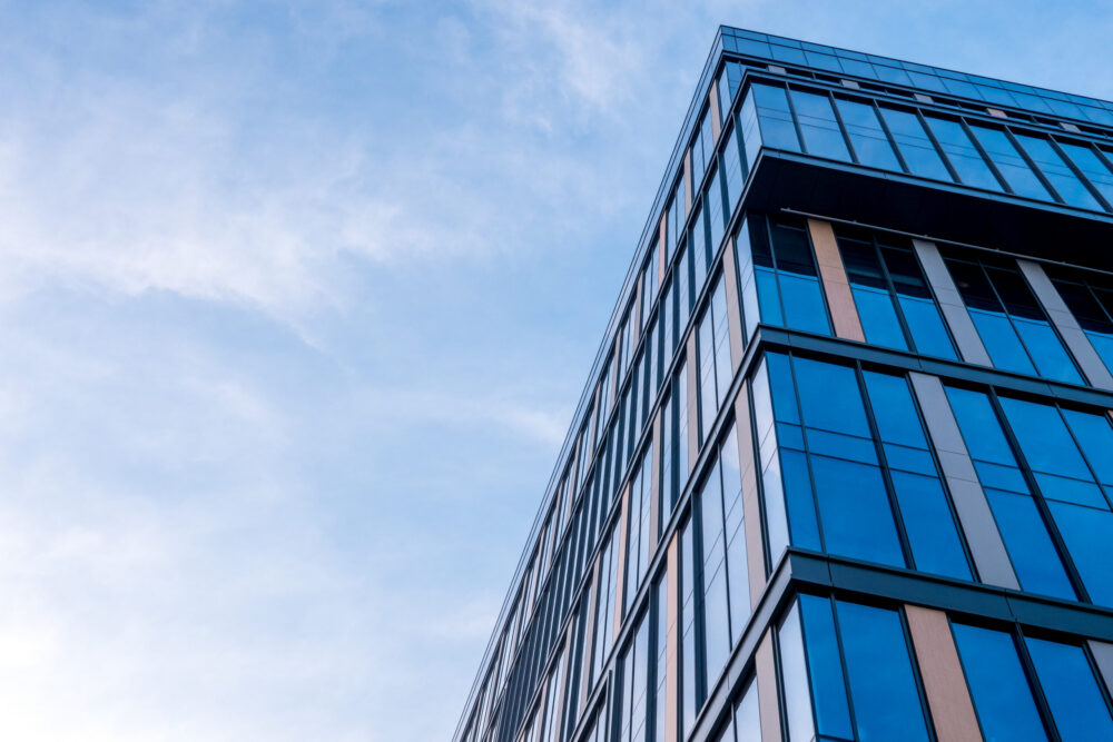 Modern glass office building viewed from a low angle against a blue sky with light clouds, showing reflective windows and sleek architectural lines.