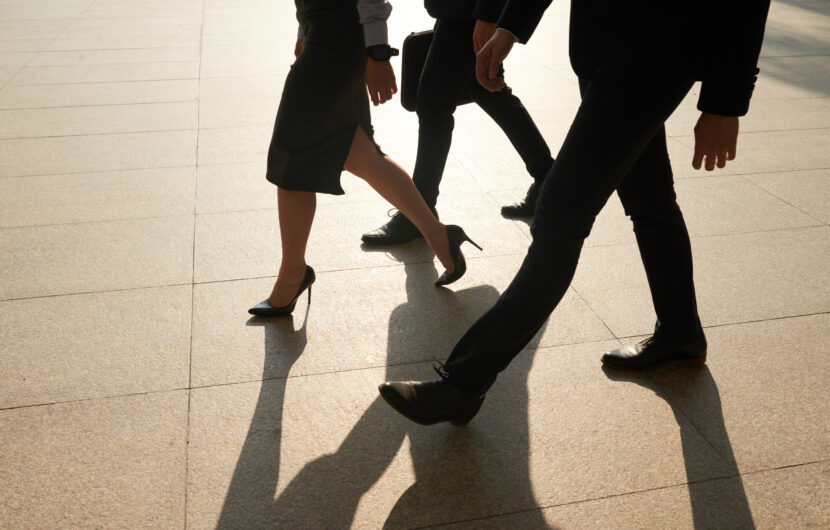 Three people dressed in business attire are walking together on a sunlit pavement, casting long shadows. Only their legs and lower bodies are visible.