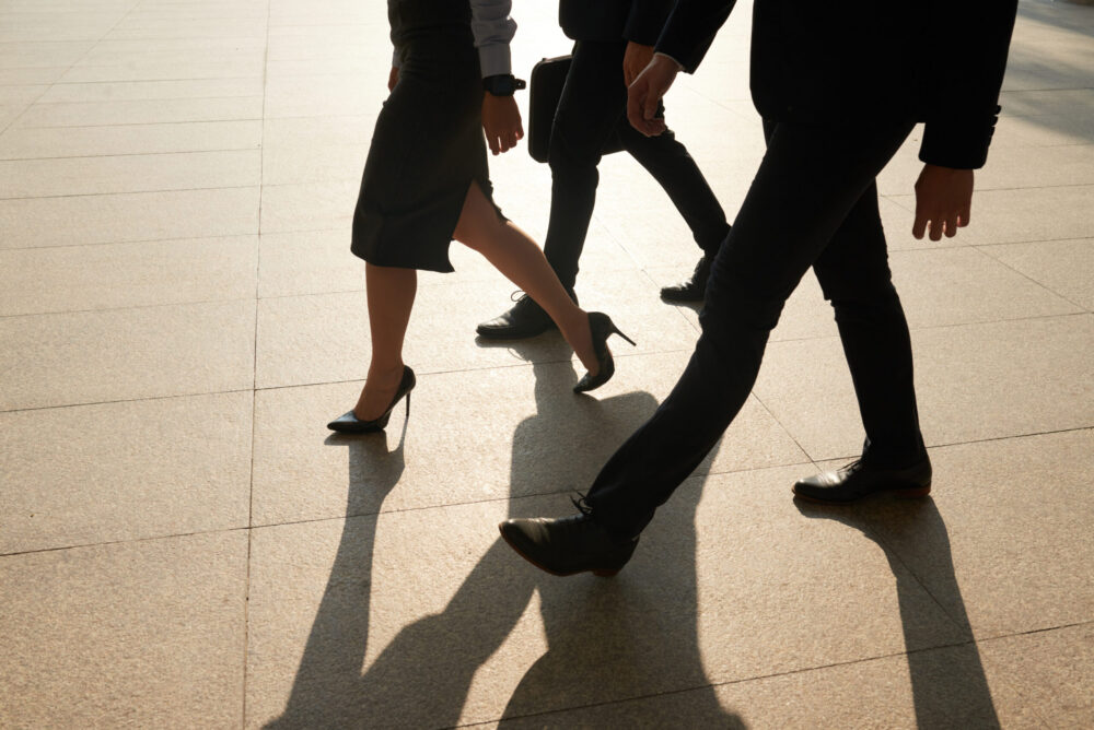 Three people dressed in business attire are walking together on a sunlit pavement, casting long shadows. Only their legs and lower bodies are visible.