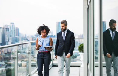 Two business professionals walk on a modern rooftop terrace with city buildings in the background. The woman is holding a tablet and talking, while the man listens attentively. Both are dressed in business attire.