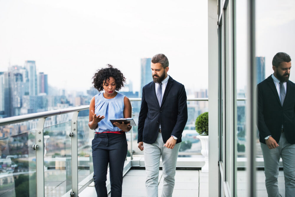 Two business professionals walk on a modern rooftop terrace with city buildings in the background. The woman is holding a tablet and talking, while the man listens attentively. Both are dressed in business attire.