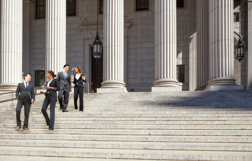 Four business professionals in suits talk and walk down the wide stone steps outside a grand building with tall columns and large hanging lanterns. The scene appears to be in front of a courthouse or government building.