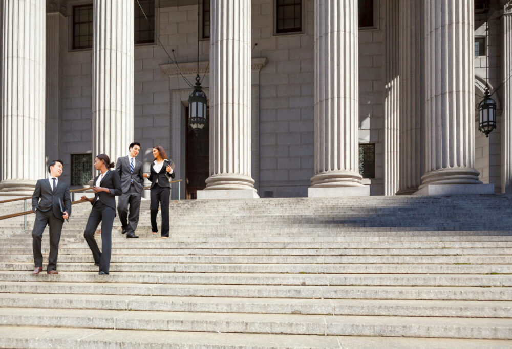 Four business professionals in suits talk and walk down the wide stone steps outside a grand building with tall columns and large hanging lanterns. The scene appears to be in front of a courthouse or government building.