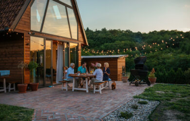 Five people sit around a table enjoying a meal on a brick patio outside a modern wooden cabin at sunset, with string lights above and a lush green forest in the background.