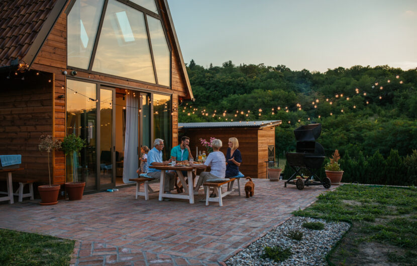 Five people sit around a table enjoying a meal on a brick patio outside a modern wooden cabin at sunset, with string lights above and a lush green forest in the background.
