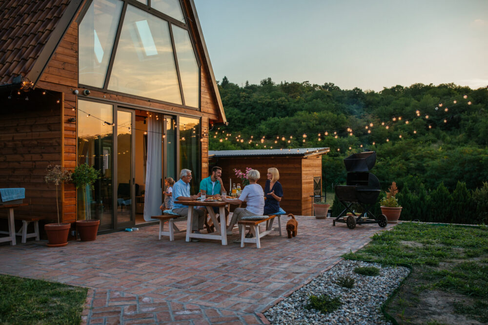 Five people sit around a table enjoying a meal on a brick patio outside a modern wooden cabin at sunset, with string lights above and a lush green forest in the background.