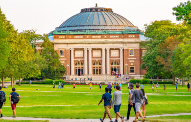 A group of students walk across a grassy quad toward a large, domed building with columns, surrounded by trees, on a university campus.