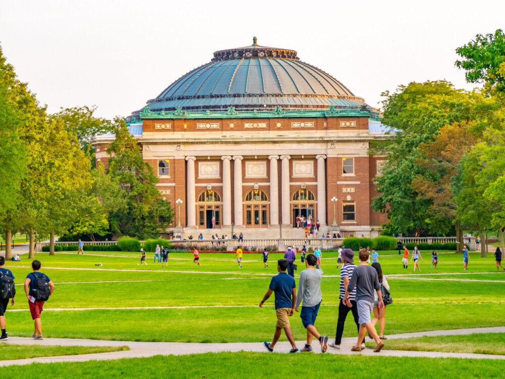 A group of students walk across a grassy quad toward a large, domed building with columns, surrounded by trees, on a university campus.